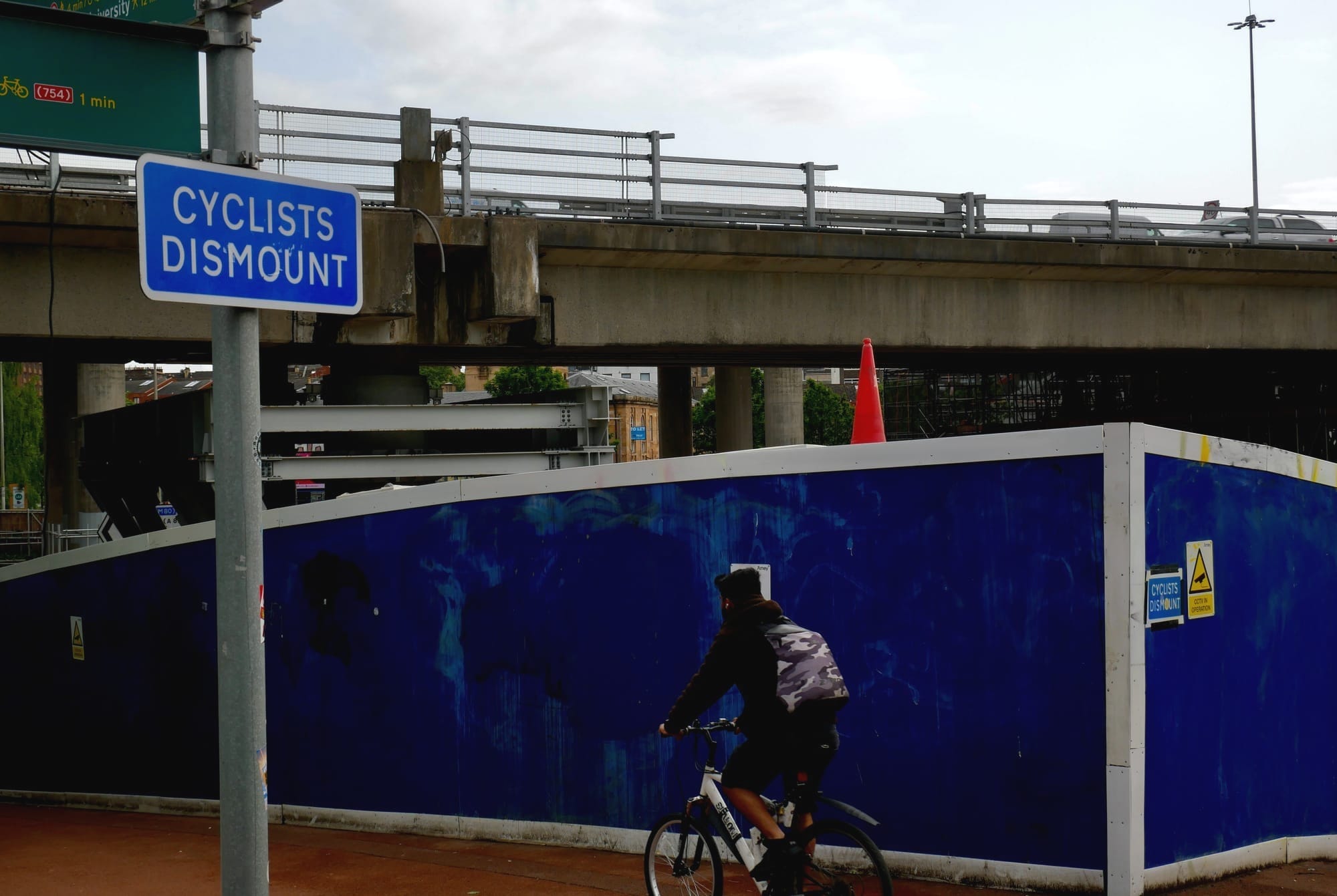 A cyclist descends on a footpath past a sign reading "cyclists dismount". The path goes under a motorway which is being partially held up by a large steel prop.