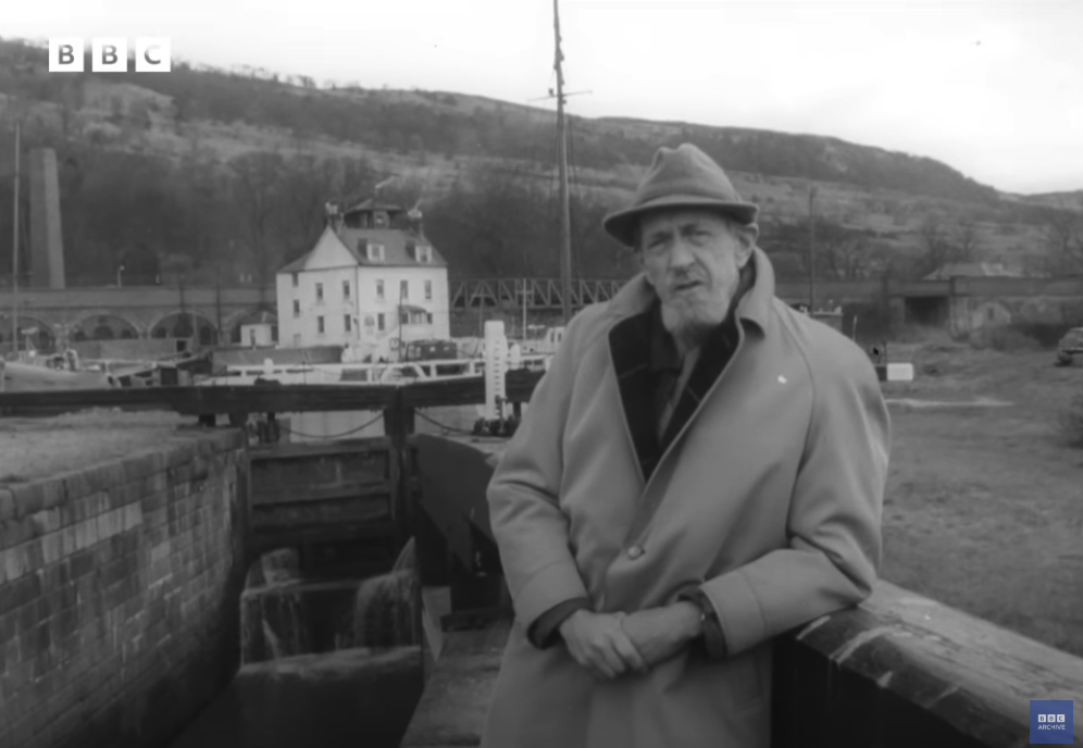 Monochrome image of an old BBC TV presenter from the 1960s. He's got one arm propped on an old wall of a canal and is wearing a large overcoat and a hat. There's a nice white house behind him.