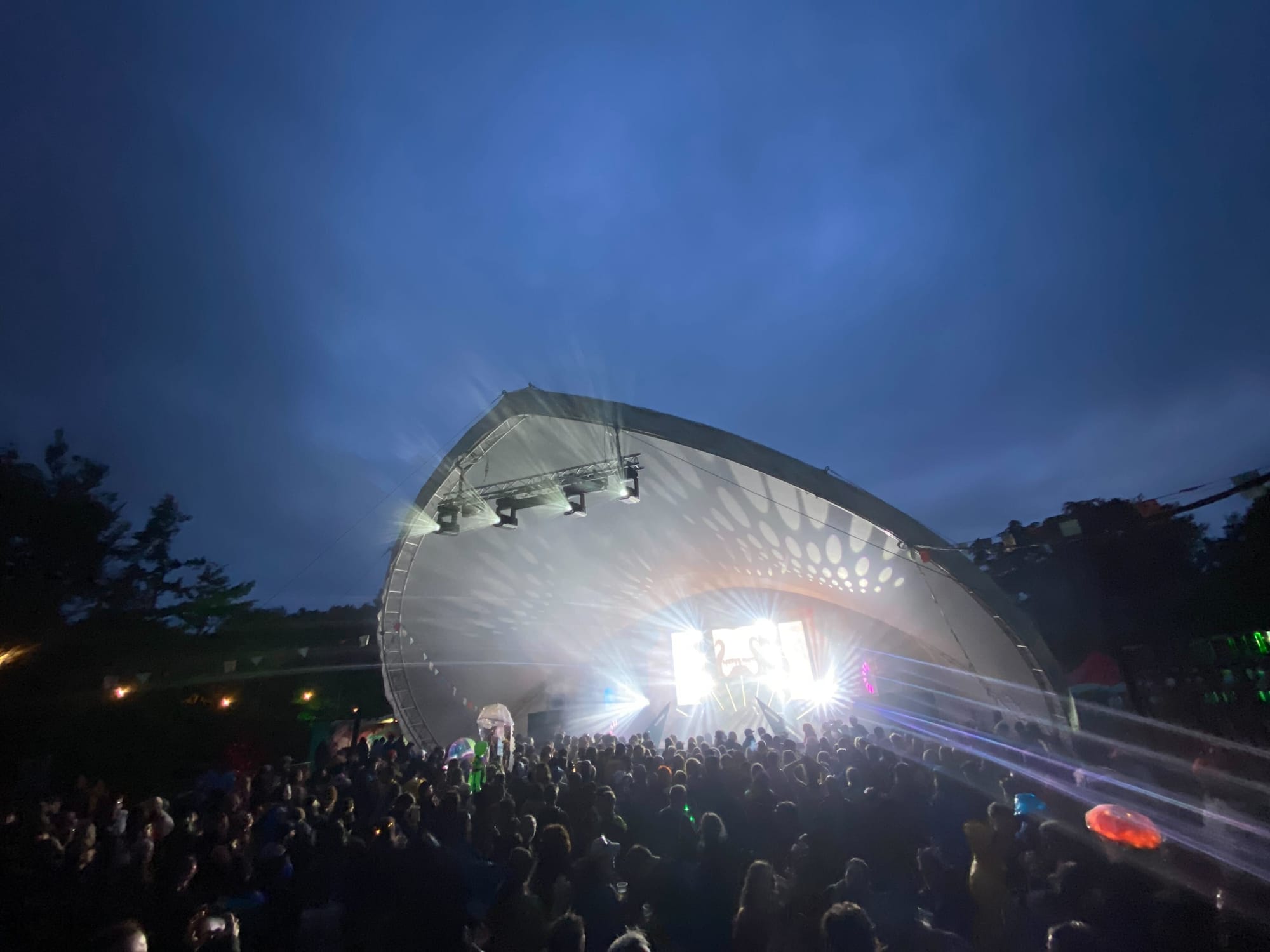An irregular-shaped stage is brightly lit, with a dark blue sky behind and a good crowd of people in front.