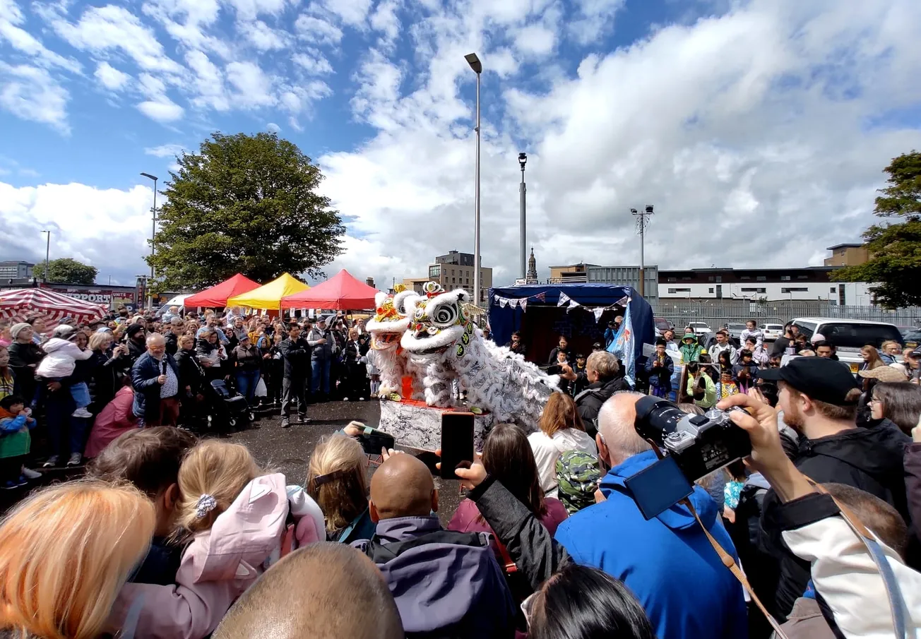 A crowd is gathered around two ornate Chinese lion costumes. Inside are two men in each with their feet on a platform. It's outside under a blue sky with fluffy clouds